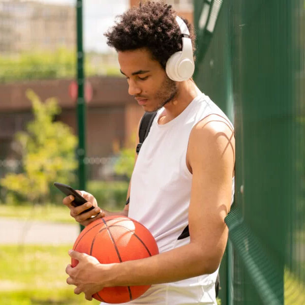 young man wearing headphones, holding a basketball, and looking at his phone